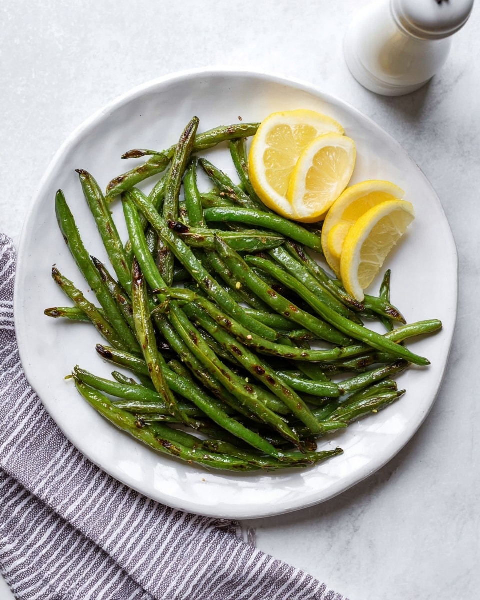 A white plate filled with a layer of cooked green beans, showing some light browning and char marks for texture, arranged in a loose pile mostly covering the plate’s center and edges; three lemon wedges placed on top of the green beans on the right side add bright yellow color contrast. The plate sits on a white marbled surface with a striped cloth partly visible on the bottom right and a white pepper grinder in the top left corner. photo taken with an iphone --ar 4:5 --v 7