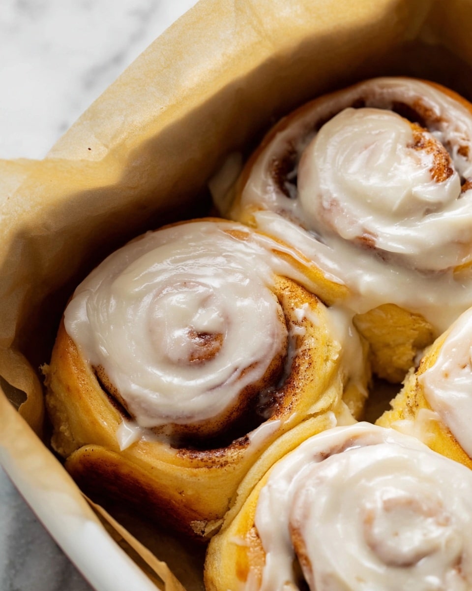 The image shows a close-up of three cinnamon rolls in a white baking dish lined with brown parchment paper. Each cinnamon roll is made of several layers of dough, golden brown and swirled with cinnamon filling, visible as darker brown lines inside the spiral. A thick layer of white cream cheese frosting covers the top of each roll, smooth with some small specks, likely vanilla or cinnamon, making a glossy texture. The background has a white marbled texture, softly blurred, putting focus on the warm, soft rolls. Photo taken with an iphone --ar 4:5 --v 7