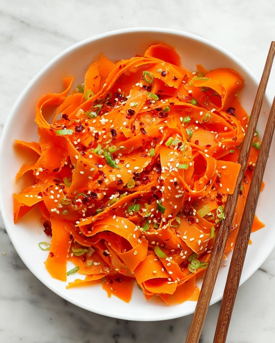 A white bowl filled with thin, wide ribbons of bright orange carrot arranged loosely in layers, sprinkled with small white sesame seeds and small bits of green onion scattered throughout. There are also tiny red chili flakes spread evenly on top. On the right side of the bowl, wooden chopsticks rest gently, partially inside the bowl. The bowl is placed on a white marbled surface. photo taken with an iphone --ar 4:5 --v 7