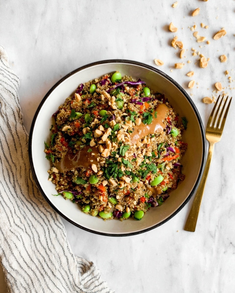 A white bowl with a black rim is filled with a colorful quinoa salad. The salad has a base layer of light brown quinoa mixed with small pieces of bright green edamame, orange-red carrot bits, and some purple cabbage pieces. On top, there are chopped green herbs and crushed peanuts scattered around, with a light brown sauce drizzled over the salad. The bowl is placed on a white marbled surface, with some crushed peanuts scattered nearby. A gold fork rests on a folded striped white cloth to the side. photo taken with an iphone --ar 4:5 --v 7