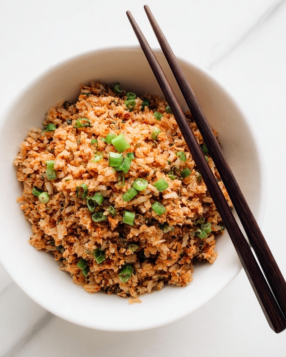A white bowl filled with a single layer of finely chopped, cooked rice mixed with small pieces of vegetables and green onion slices scattered on top, creating a mix of light brown and green colors with a slightly crumbly texture. Two dark wooden chopsticks rest diagonally on the bowl's edge. The bowl is placed on a white marbled surface. photo taken with an iphone --ar 4:5 --v 7