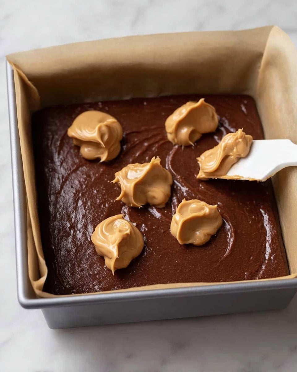 A square gray baking pan lined with parchment paper is shown on a white marbled surface. Inside the pan is a thick, smooth dark brown chocolate batter evenly spread to cover the base. On top, there are four dollops of creamy light brown peanut butter, three already placed and one being added with a white spatula. The peanut butter dollops have a soft, glossy texture and stand out against the matte chocolate layer. photo taken with an iphone --ar 4:5 --v 7