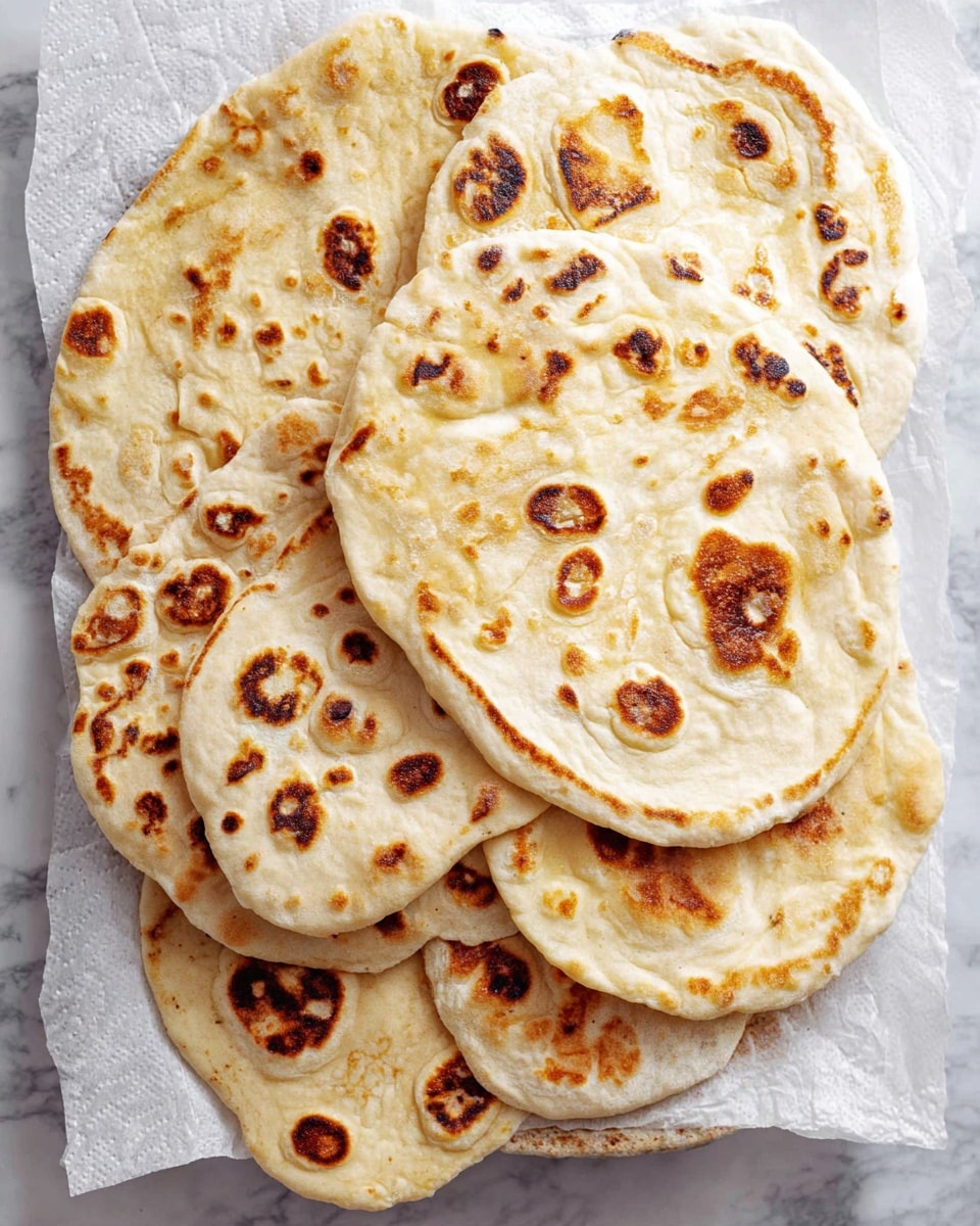 A stack of seven flatbreads with golden brown spots and slightly puffed textures is arranged overlapping each other on white parchment paper. The flatbreads have uneven round shapes with some areas showing darker char marks, creating a contrast with the lighter off-white dough. The flatbreads look soft and slightly thick, showing air bubbles and textured surface with spots spread irregularly. The whole presentation sits on a white marbled surface. photo taken with an iphone --ar 4:5 --v 7