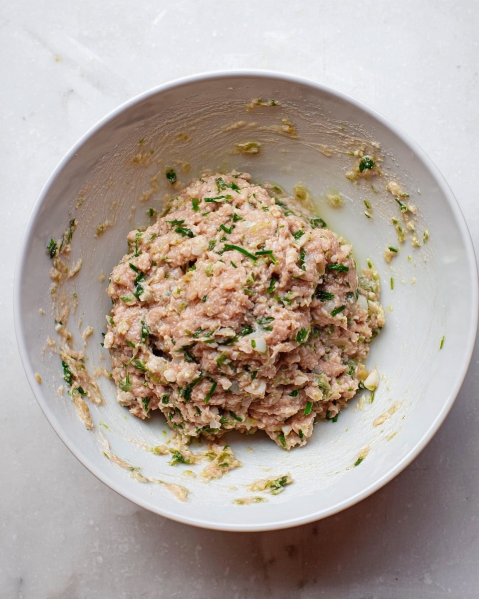Inside a white bowl, there is one layer of raw minced mixture that looks soft and slightly sticky, with a pale pink color mixed with green bits of herbs and small white pieces. The mixture sits loosely in the middle of the bowl, with some of it spread thinner along the sides, showing a rough texture with visible small chunks. The bowl rests on a white marbled surface, and the lighting softly highlights the moist, fresh look of the mixture photo taken with an iphone --ar 4:5 --v 7