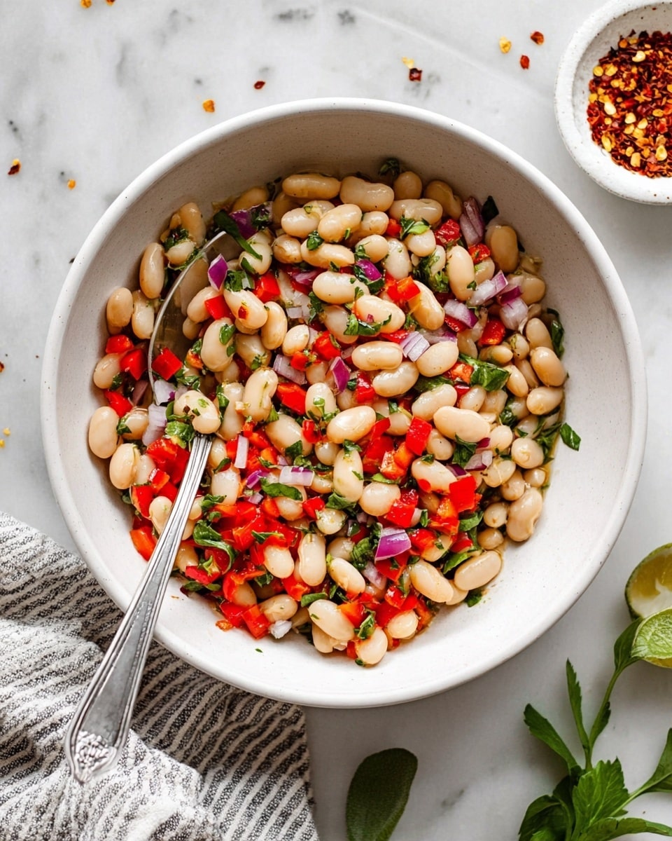 A white bowl filled with a colorful bean salad showing three main layers: the bottom layer is white beans, the middle layer consists of diced red bell peppers and small pieces of red onion, and the top layer has scattered green herb leaves mixed throughout. A silver spoon rests inside the bowl with some beans on it. Around the bowl, there is a white marbled surface with a striped cloth partially visible at the bottom right, a small white dish with red pepper flakes on the top right, and some green herb leaves on the sides. photo taken with an iphone --ar 4:5 --v 7