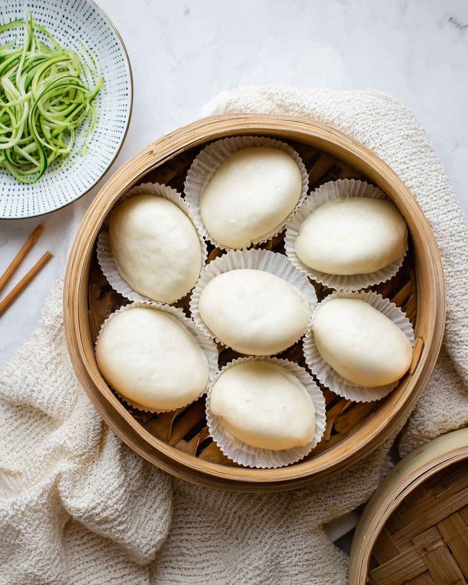 A round bamboo steamer basket filled with two layers of white, soft, oval-shaped steamed buns, each resting on small, round white paper liners. The buns are smooth and slightly puffy, arranged close together to fill the basket. The basket sits on a white marbled surface with a light cream-colored knit cloth underneath part of it. In the top left corner, there is a small white plate with blue dots holding thin green spirals of vegetable, next to a pair of wooden chopsticks resting on the marbled surface. photo taken with an iphone --ar 4:5 --v 7