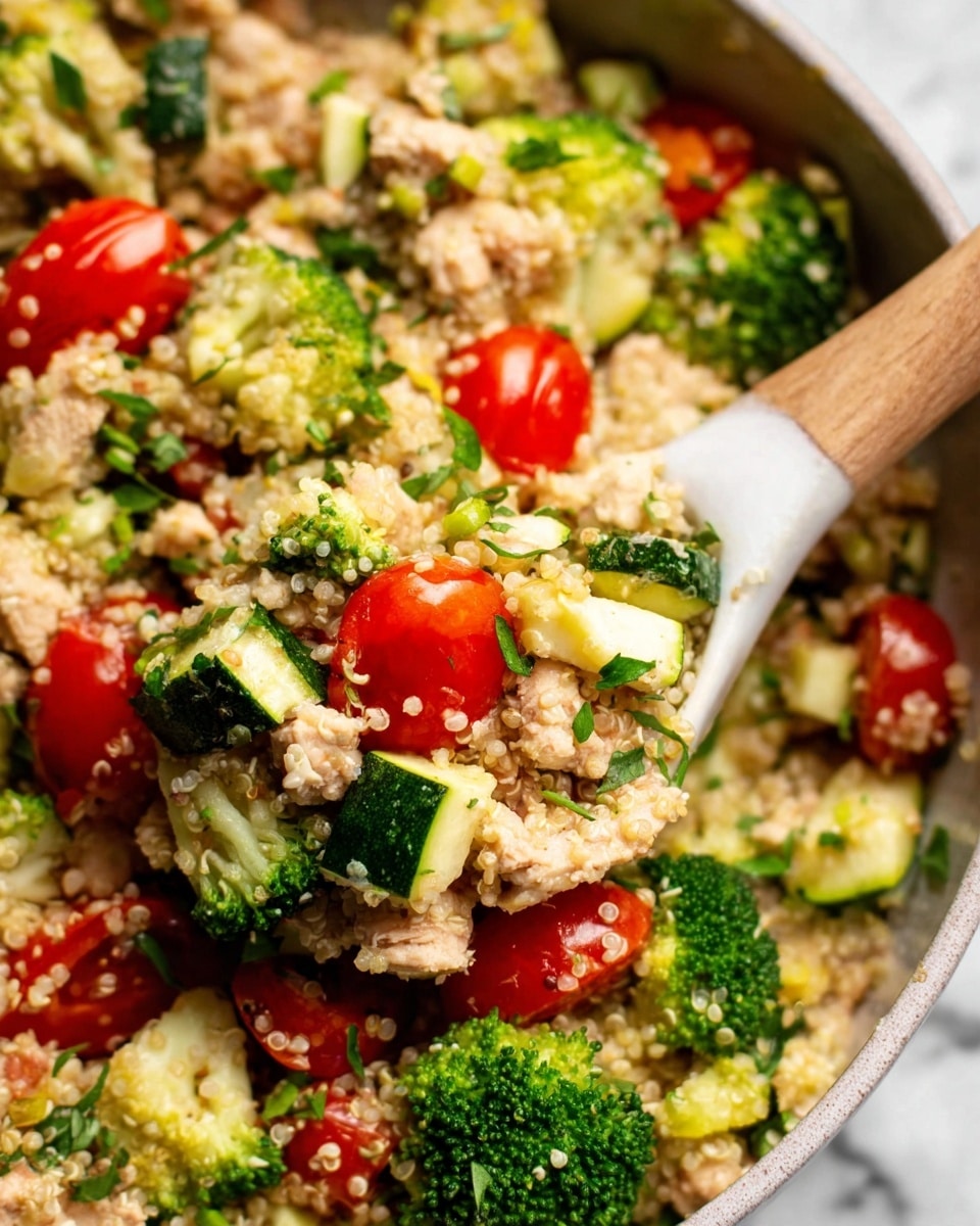 The image shows a close-up of a mixed salad with three main layers. The bottom layer is small, light beige quinoa grains with a soft, grainy texture. Above that are bright green broccoli florets and zucchini pieces, chopped into small chunks, adding a fresh, rough texture. On top, there are bright red cherry tomatoes, some whole and some sliced in halves, showing smooth shiny skin and juicy interiors, mixed with small bits of cooked chicken in light beige color, and scattered fresh green herbs for garnish. A white spoon with a wooden handle is scooping a portion of this colorful salad. The dish is set against a white marbled surface. photo taken with an iphone --ar 4:5 --v 7