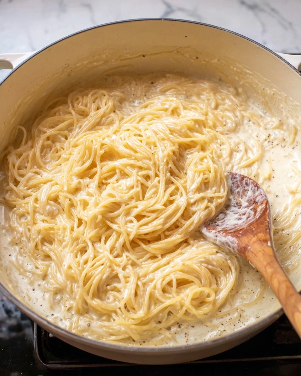 In a white pot, there is a creamy layer of thin spaghetti noodles evenly coated in a pale yellow sauce with a smooth, slightly bubbly texture, giving it a rich and creamy appearance. The noodles are softly piled in the center with some strands loosely spread around. On the right, a wooden spoon with traces of sauce rests inside the pot, its surface showing a mix of the sauce’s creaminess and slightly darker spots from seasoning. The whole scene is set against a white marbled background. photo taken with an iphone --ar 4:5 --v 7