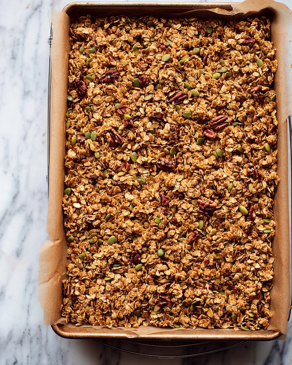 A large flat layer of golden brown granola spread evenly on a baking sheet lined with brown parchment paper, showing a mix of rolled oats, green pumpkin seeds, and scattered whole pecans adding texture and darker brown color contrast throughout. The baking sheet rests on a wire rack over a white marbled surface, with the granola’s texture appearing crunchy and loose, revealing its natural ingredients in a close-up view. Photo taken with an iphone --ar 4:5 --v 7