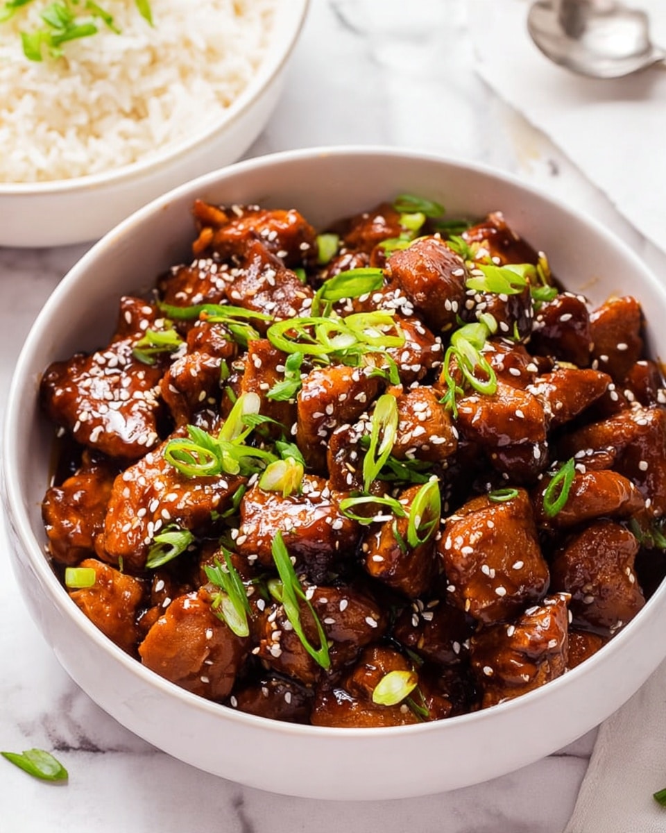 A white bowl filled with shiny, dark brown chunks of chicken coated in a thick sauce, topped with white sesame seeds and bright green sliced scallions scattered evenly over the top. The chicken pieces are bite-sized, layered closely together, and glisten under soft lighting. The bowl sits on a white marbled surface, with a partial view of another white bowl containing plain white rice garnished with a few small green scallion pieces. A silver spoon is partially visible to the side. Photo taken with an iphone --ar 4:5 --v 7