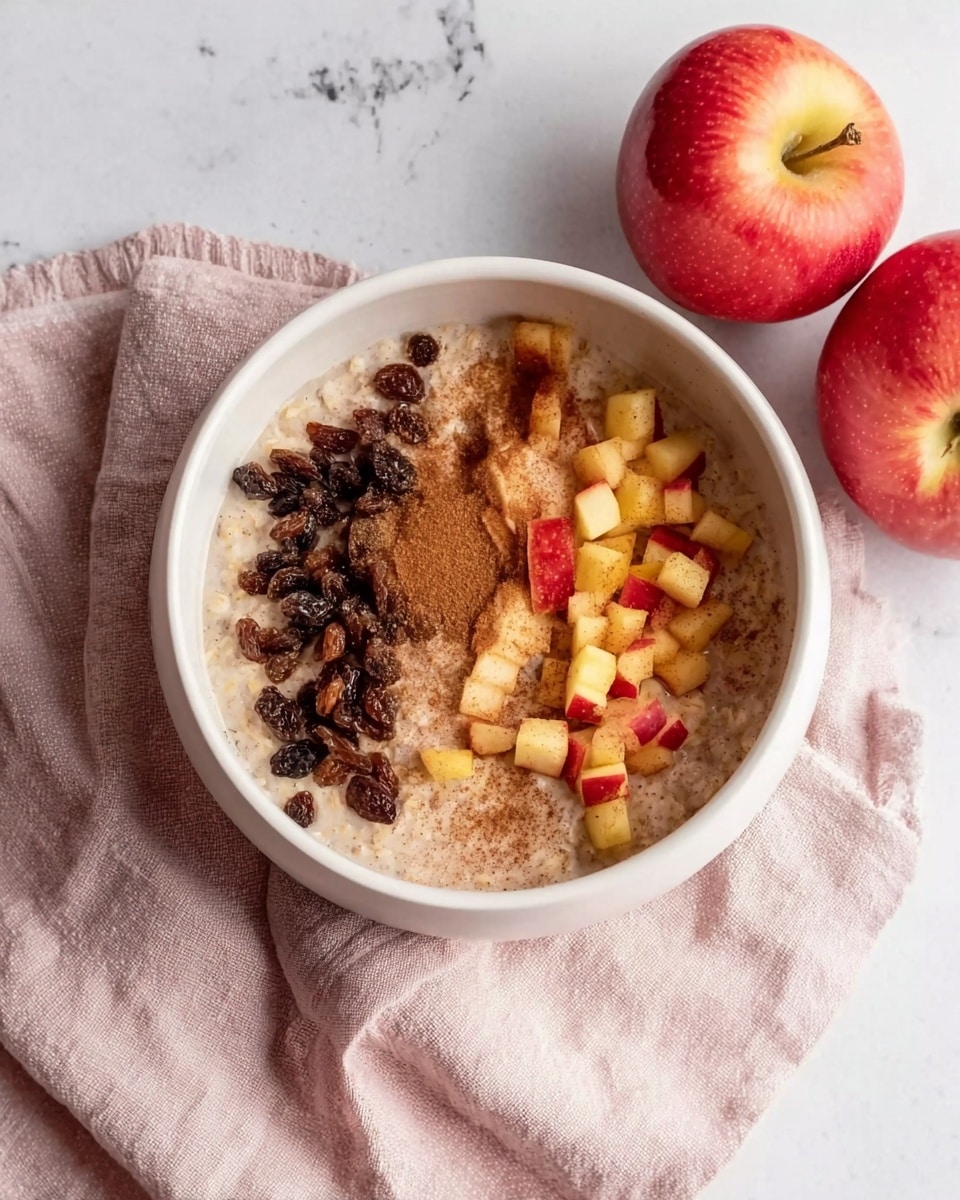 The image shows a white bowl filled with a light beige oatmeal base. On top of the oatmeal, there are two separate piles of toppings: dark brown raisins on the left side and small diced pieces of red and yellow apple on the bottom right. The oatmeal surface has a light sprinkle of cinnamon powder on the right side. The bowl is placed on a soft pink and white cloth, all set on a white marbled surface. Two fresh red apples are placed nearby on the marbled surface. photo taken with an iphone --ar 4:5 --v 7