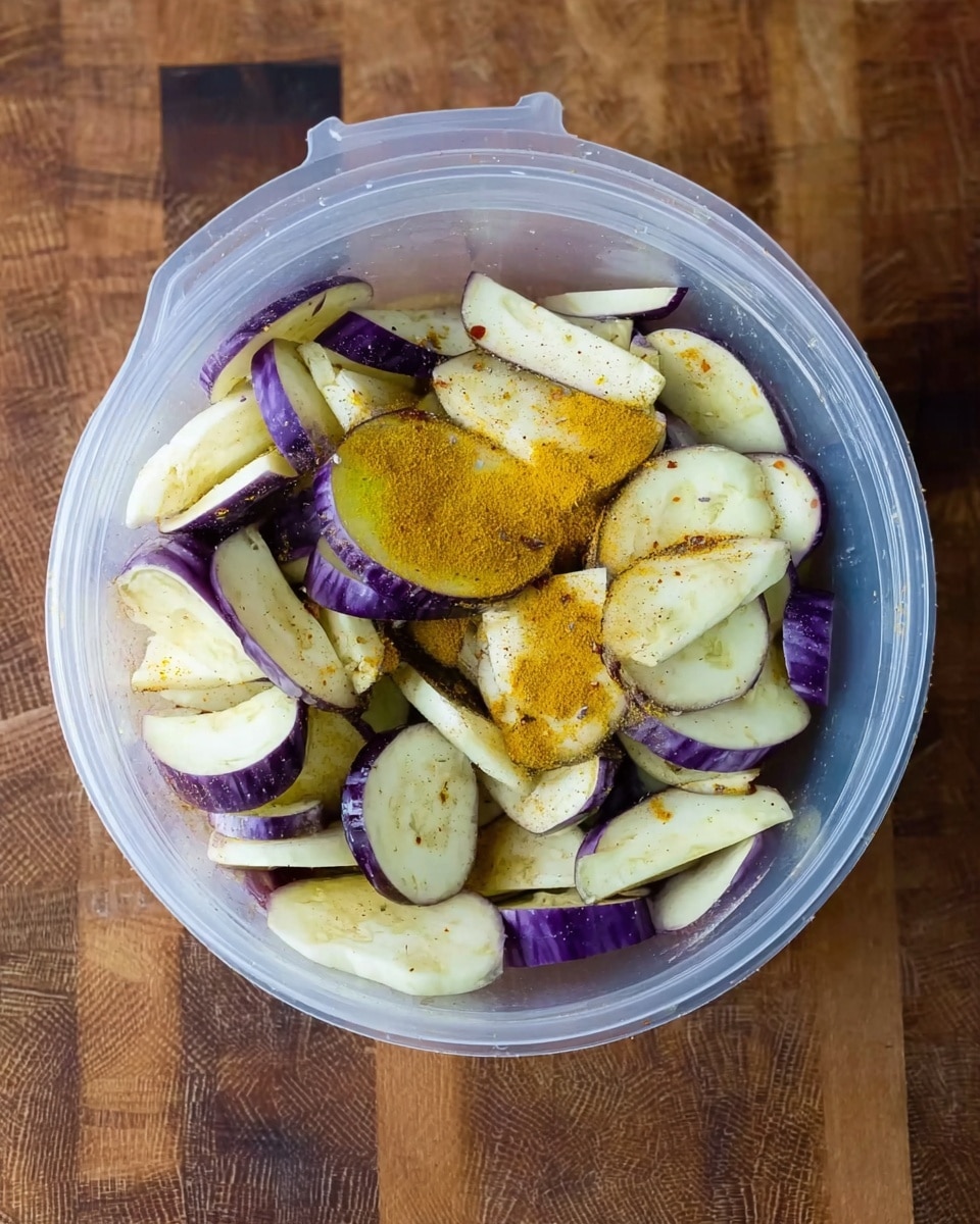 A clear plastic container filled with many slices of eggplant showing white and purple colors, with some light brown spots on the white parts. A bright yellow powder, likely spice, is sprinkled on top of the eggplant slices, mostly in the center. The container is on a wooden surface with a textured pattern. photo taken with an iphone --ar 4:5 --v 7