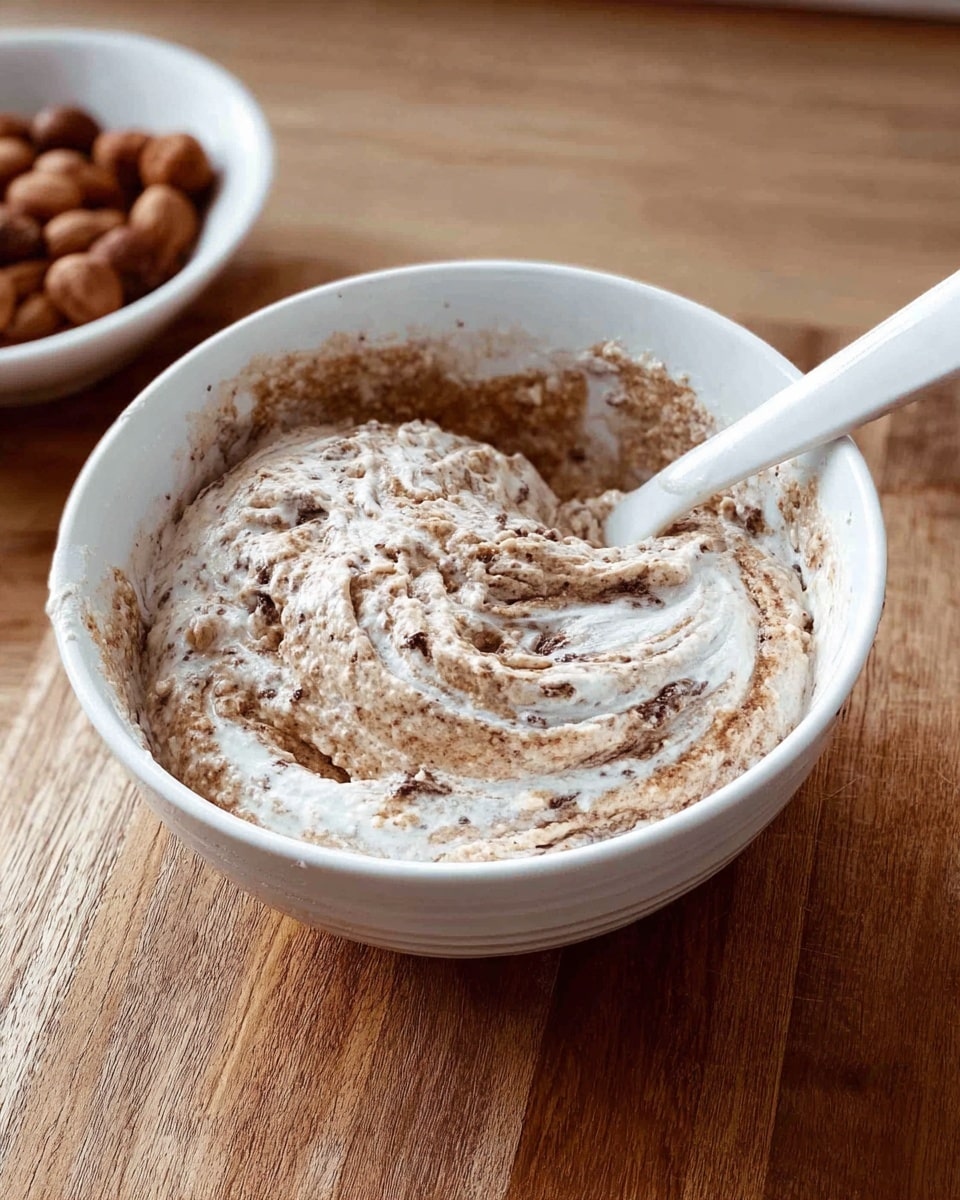 A white bowl sits on a wooden surface filled with a mixture that has two main layers: a thick, creamy white base with a rough, grainy brown layer swirled throughout and on top, giving a marbled effect. A white spoon is partially inside the bowl, resting on the side, covered with the same mixture. In the background, to the left, there is a partially visible bowl filled with round brown nuts. photo taken with an iphone --ar 4:5 --v 7