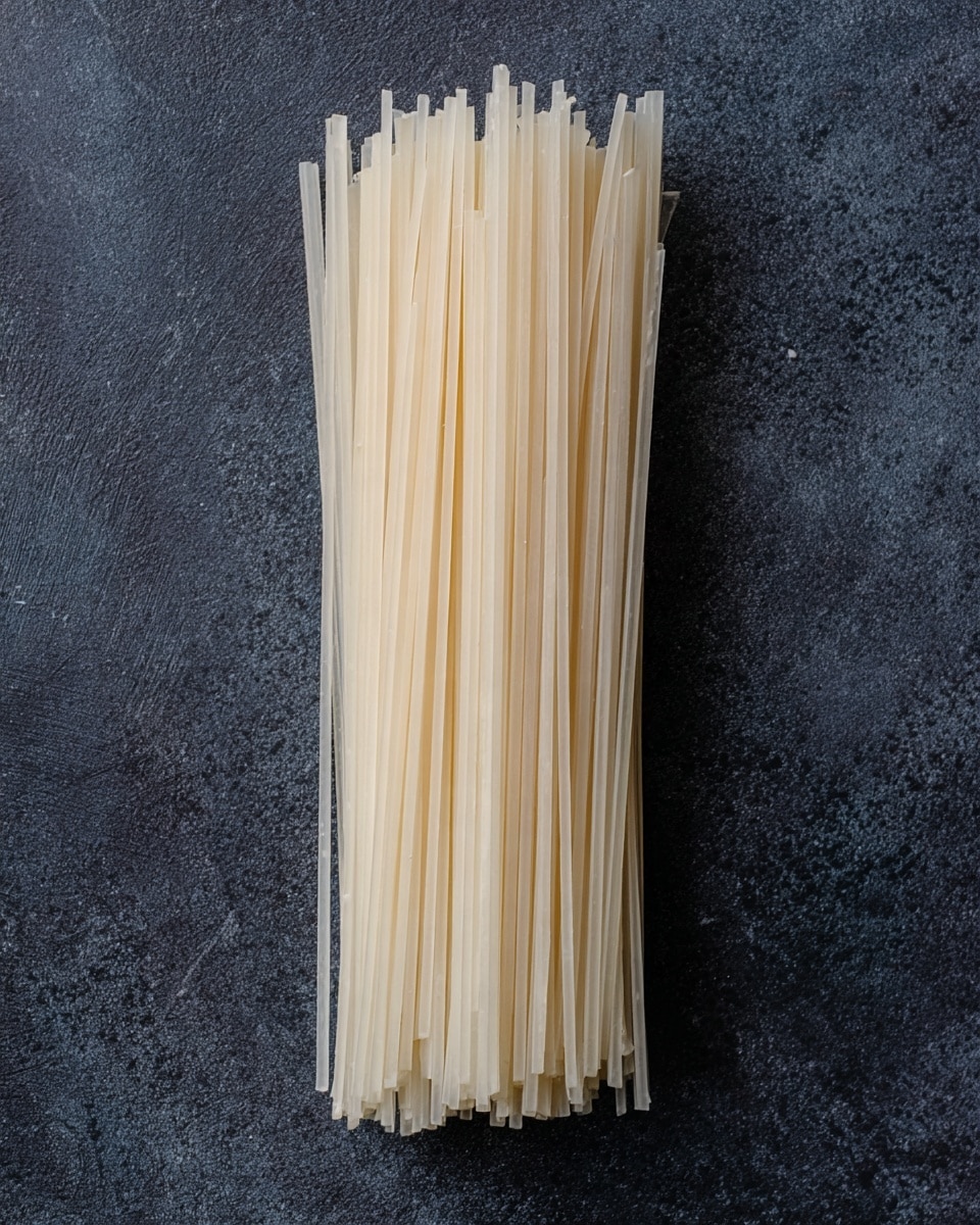 A bundle of uncooked flat rice noodles is laid out vertically on a dark gray textured surface, each noodle thin and slightly translucent with a pale off-white color. The noodles are neatly stacked together in a compact, slightly uneven layer that shows small variations in noodle width and length at the edges. The surface underneath looks like dark stone with a rough, natural texture. photo taken with an iphone --ar 4:5 --v 7