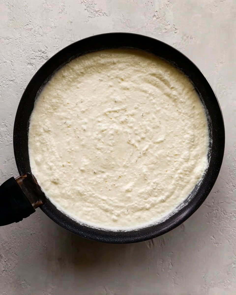 A round black pan filled with a thick creamy white mixture that covers the entire pan surface evenly. The mixture has a smooth texture with small bits visible inside, creating a slightly lumpy look across the top. The pan handle extends outwards on the left side and the pan rests on a white marbled surface. Photo taken with an iphone --ar 4:5 --v 7