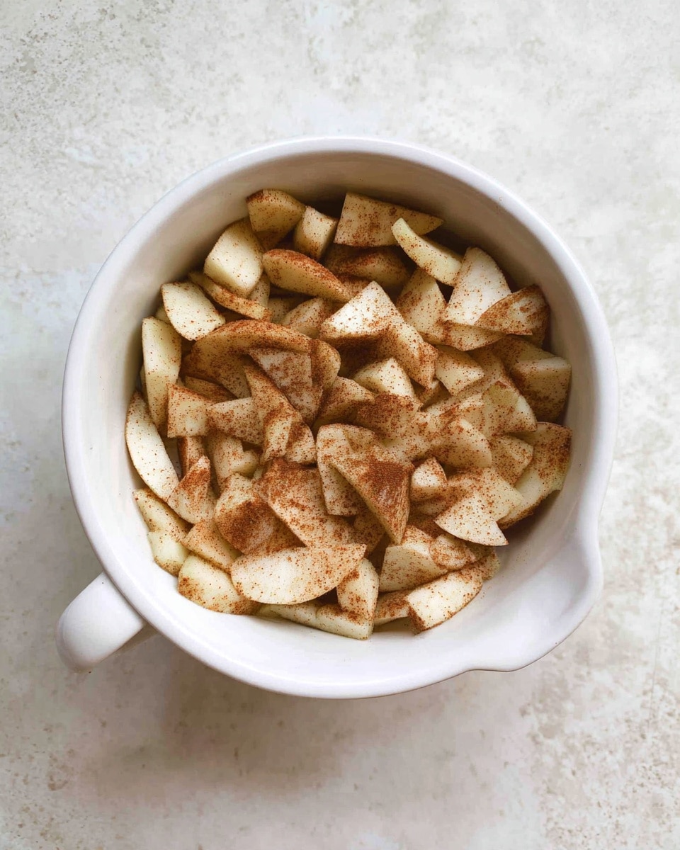 A white bowl with a handle contains many small pieces of peeled apple slices covered evenly with a light brown cinnamon powder, giving a speckled texture to the pale apple pieces. The bowl is sitting on a white marbled surface. photo taken with an iphone --ar 4:5 --v 7