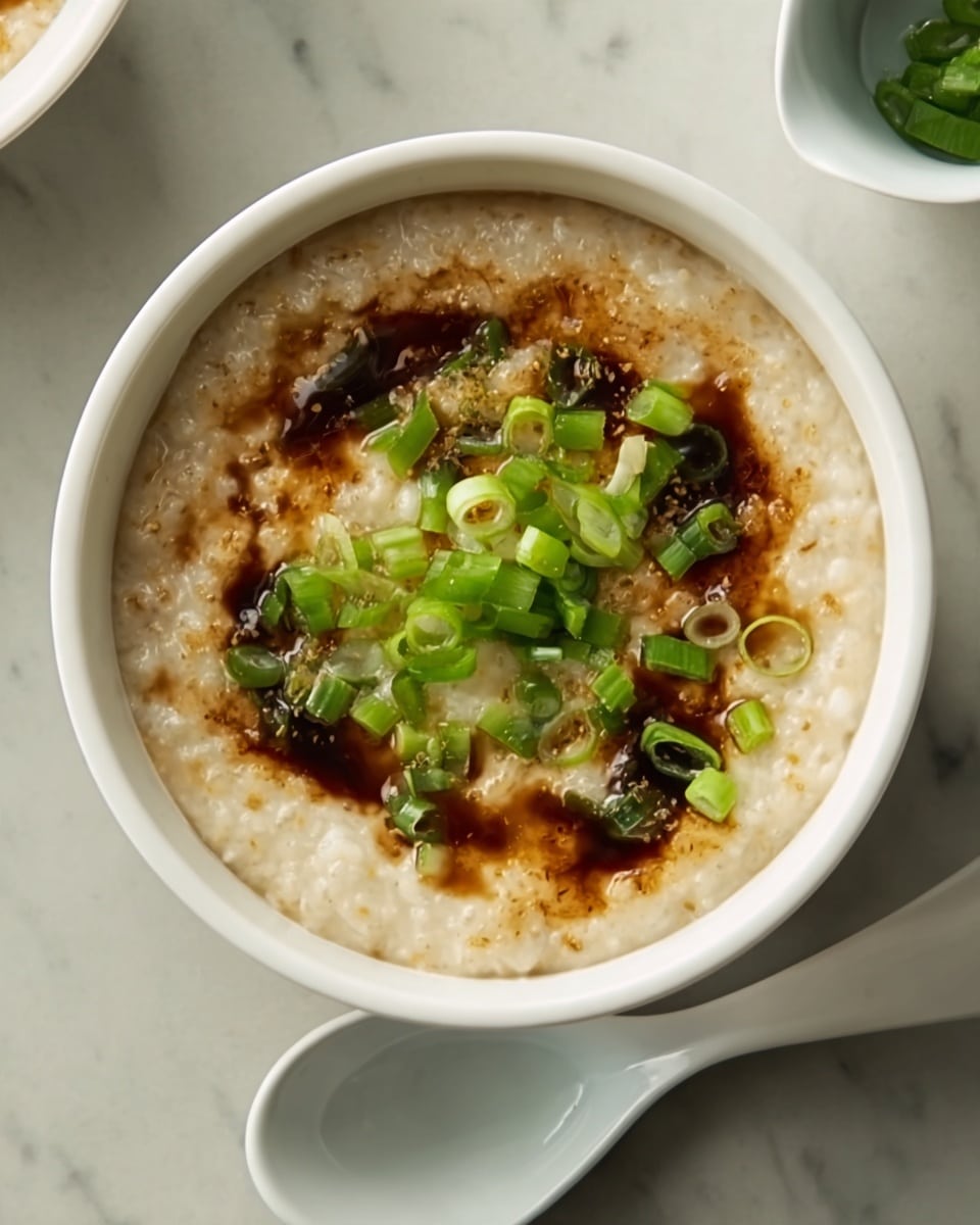 A white bowl filled with a thick, creamy porridge that has a light beige color. On top, there is a layer of dark brown sauce spread unevenly, adding contrast. Green chopped scallions are sprinkled in the center, providing a fresh, bright green color and a bit of texture to the smooth porridge base. The bowl is placed on a white marbled surface next to a white spoon, highlighting the colors of the dish. Photo taken with an iphone --ar 4:5 --v 7
