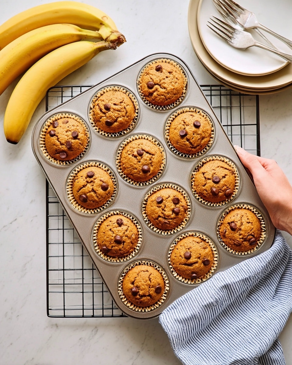 A 12-slot muffin pan holds twelve golden brown muffins with small chocolate chips visible on the cracked tops, each muffin sitting in a light tan paper liner. The pan rests on a wire cooling rack above a white marbled surface. To the top left, two yellow bananas with some brown spots curve gently. At the top right, a stack of white plates has two forks resting on the top plate. A woman's hand with light skin holds a blue and white striped kitchen towel, grasping the bottom right corner of the muffin pan. photo taken with an iphone --ar 4:5 --v 7