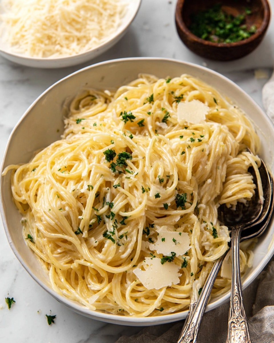 A large white bowl filled with creamy spaghetti pasta that looks soft and smooth, coated evenly with a light yellow sauce. On top are small green parsley pieces and thin, pale yellow cheese shavings scattered over the noodles. Two vintage silver spoons rest on the side of the bowl, one holding a small bundle of twisted spaghetti. In the background, there is a white bowl filled with more cheese shavings and a small dark brown bowl with green herbs, all set on a white marbled surface. Photo taken with an iphone --ar 4:5 --v 7