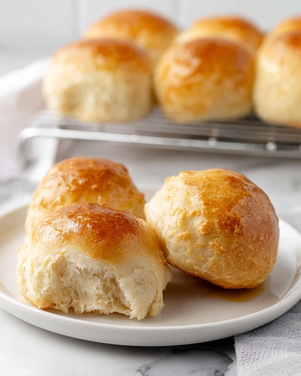 The image shows a close-up of a white plate holding four soft bread rolls with a shiny, golden-brown top crust and a light, fluffy inside texture. In the background, more bread rolls are cooling on a metal wire rack, with a white marbled countertop beneath and a light gray cloth to the side. The soft bread rolls have uneven, rounded shapes with a slightly crumbly surface on the sides, and the shiny tops catch the light, showing a smooth texture. photo taken with an iphone --ar 4:5 --v 7